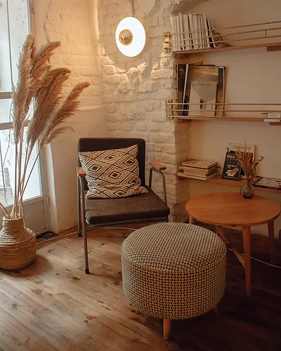 A cushioned chair, a round footstool and a small wooden table in a softly lit corner with shelves of books.